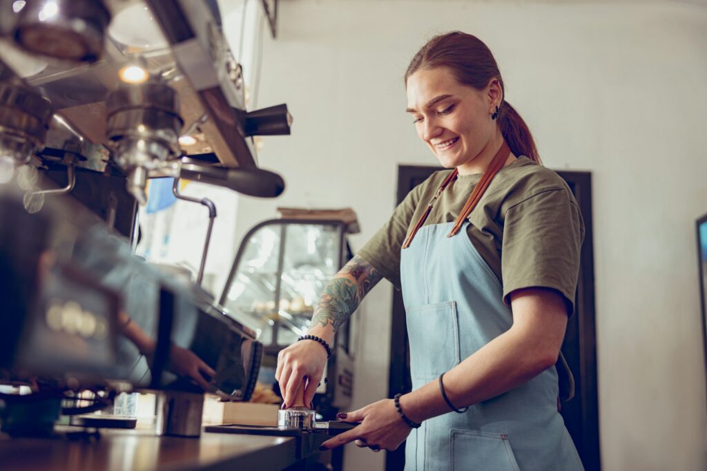 Cheerful woman barista making coffee in cafeteria